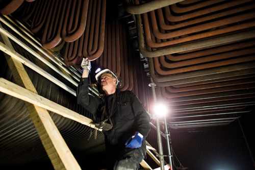 Inspektör i skyddsutrustning kontrollerar metallrör i en industriell anläggning, med byggnadsställningar och belysning i närheten.