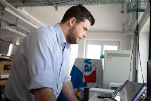 Kiwa lab employee in a laboratory setting focusing intently on a computer screen, wearing a light blue shirt; Lab filled with technical equipment