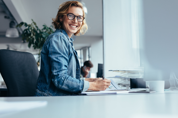 Smiling woman in a denim jacket working at an office desk with a notepad and pen