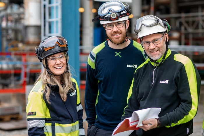 Three Kiwa inspectors wearing safety gear and helmets smiling at a work site, with one holding documents