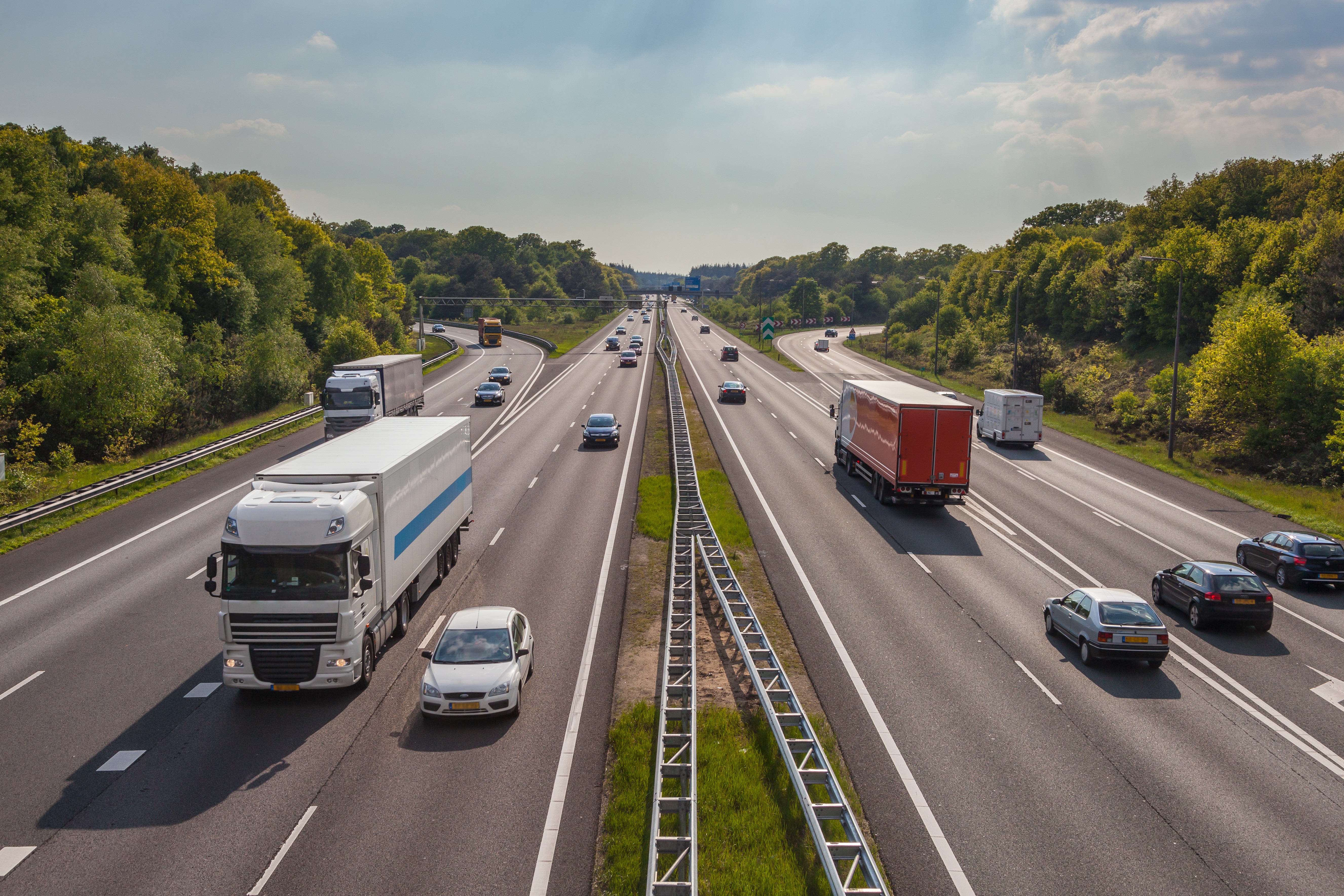 Highway view with cars and trucks surrounded by lush green trees under a partly cloudy sky.