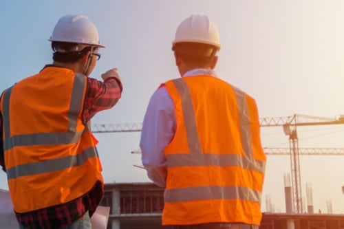 Two Kiwa auditors in orange safety vests and helmets surveying a building site with cranes in the background under a clear sky