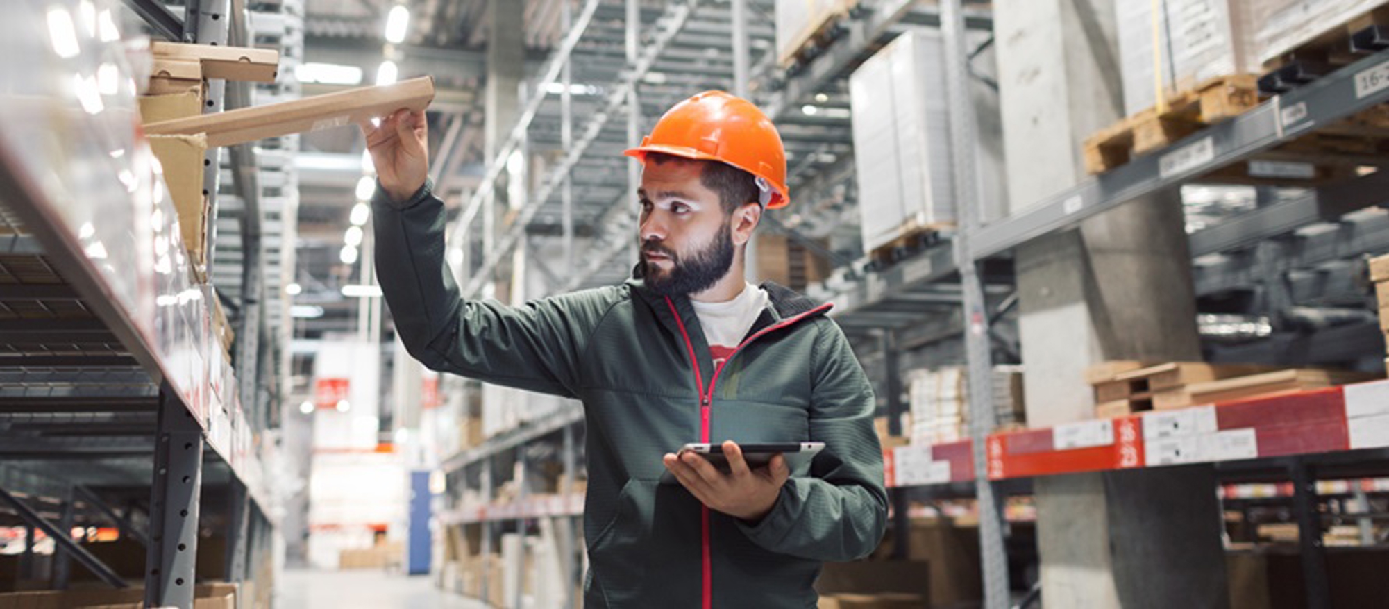 Warehouse worker wearing an orange hard hat inspects inventory on shelves while using a tablet in a large industrial storage facility.