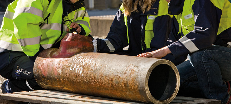Ingenjörer i varselkläder inspekterar ett stort rostigt metallrör som ligger på en träpall.