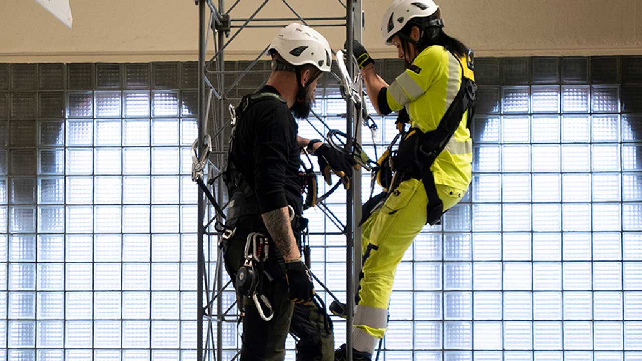 Två personer med säkerhetsutrustning klättrar på en metallstruktur, iklädda skyddshjälmar och arbetskläder.