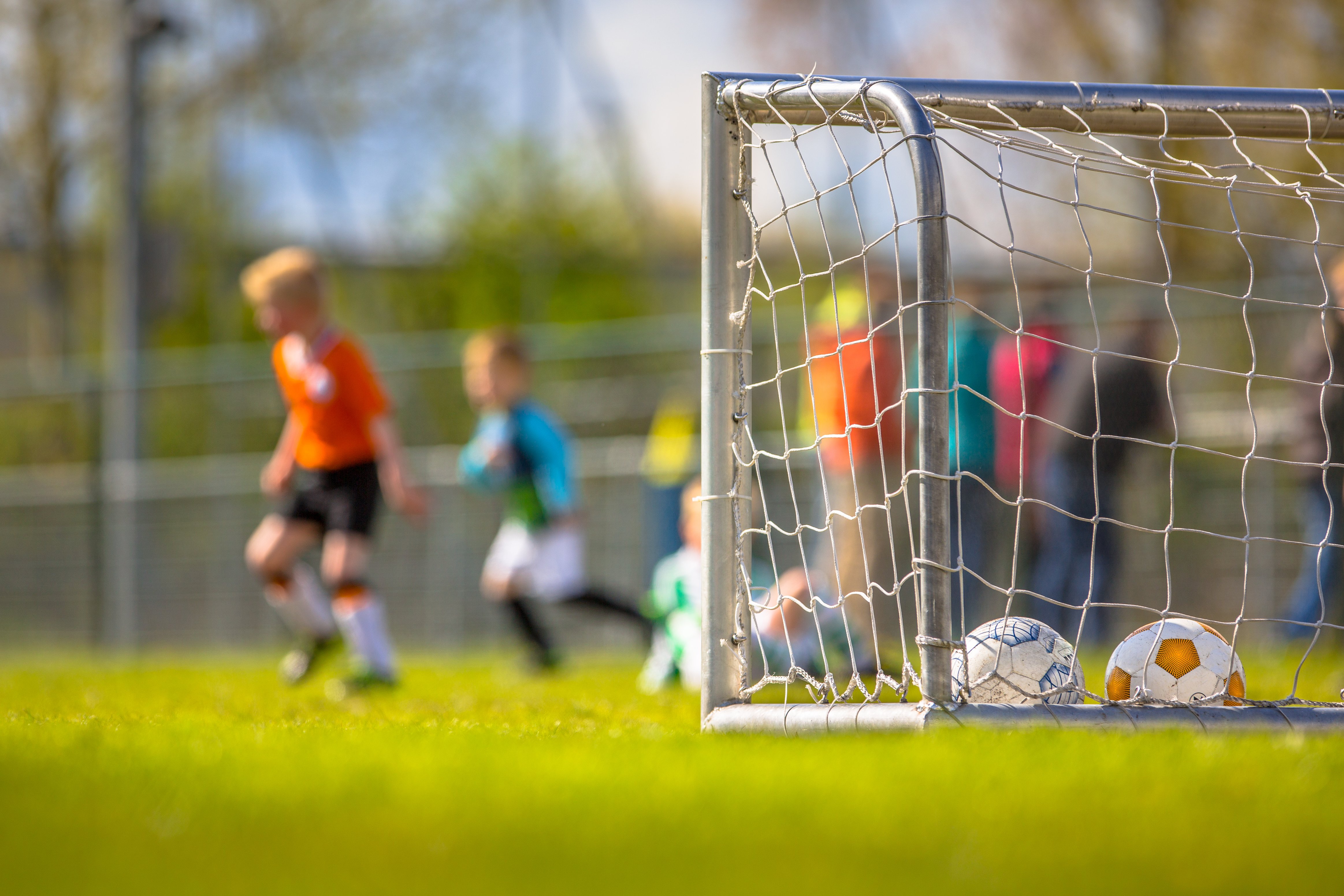 A soccer goal with two balls inside on a grassy field, with children playing soccer in the background.