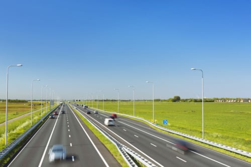 Autos rijden op een snelweg met meerdere rijstroken omgeven door groene weilanden onder een blauwe lucht.