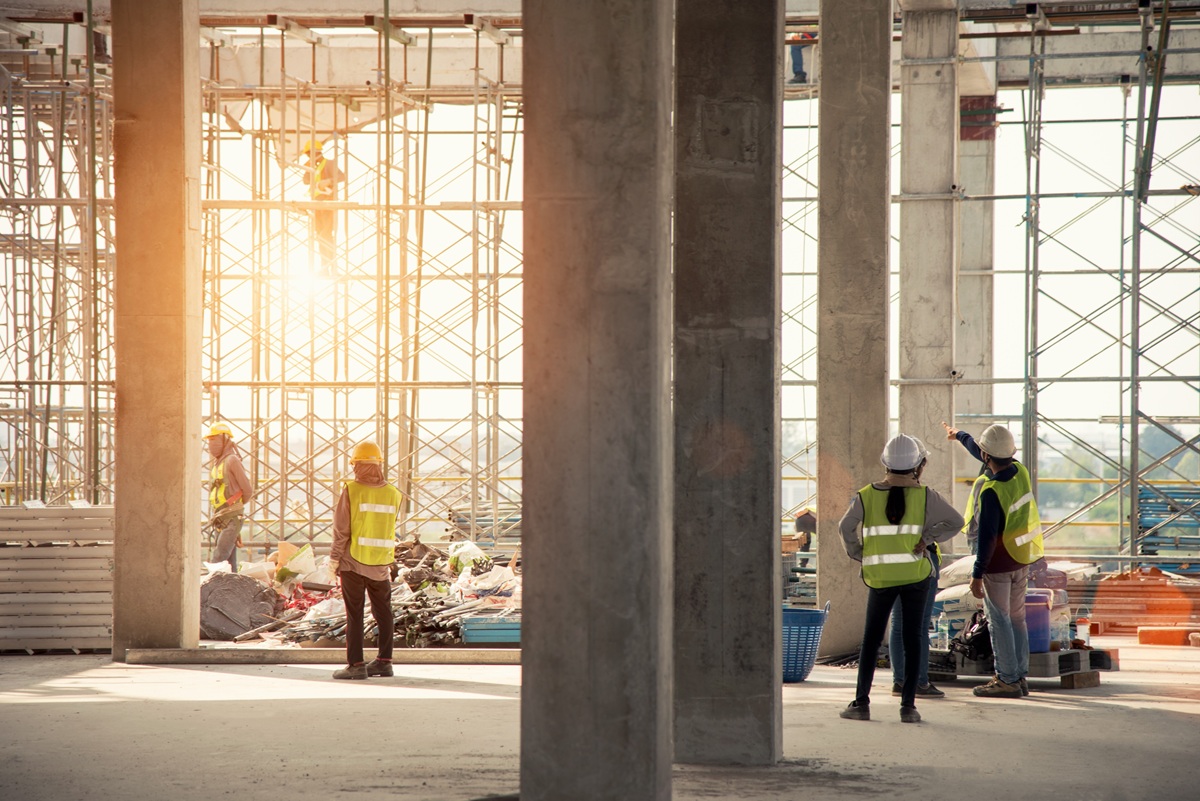 Bouwplaats in een constructiehal met steigers, betonnen kolommen en meerdere bouwvakkers met gele veiligheidshesjes en helmen bij zonlicht.
