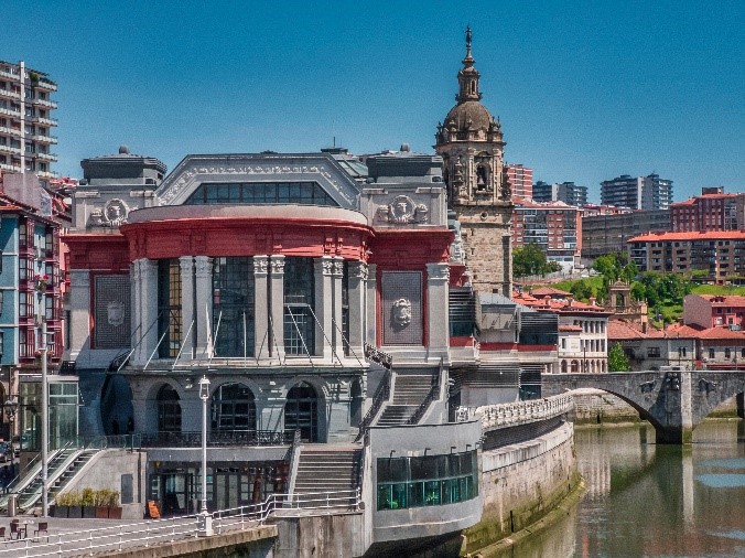 Vista urbana de un edificio histórico junto a un río, con una torre de iglesia al fondo y puentes de piedra sobre el agua.