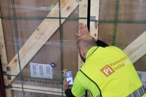 Trabajador inspeccionando un módulo solar embalado en una caja de madera, utilizando un dispositivo móvil, con chaleco de seguridad del Instituto Fotovoltaico.