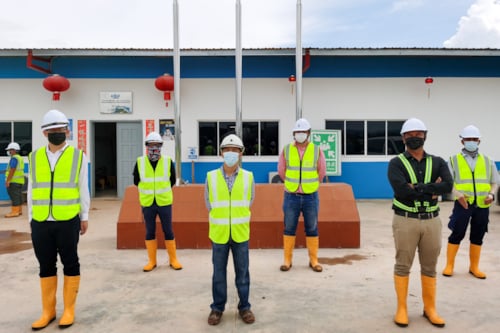 Trabajadores de la construcción con cascos y chalecos reflectantes amarillos, de pie frente a un edificio de obra con botas de seguridad naranjas.