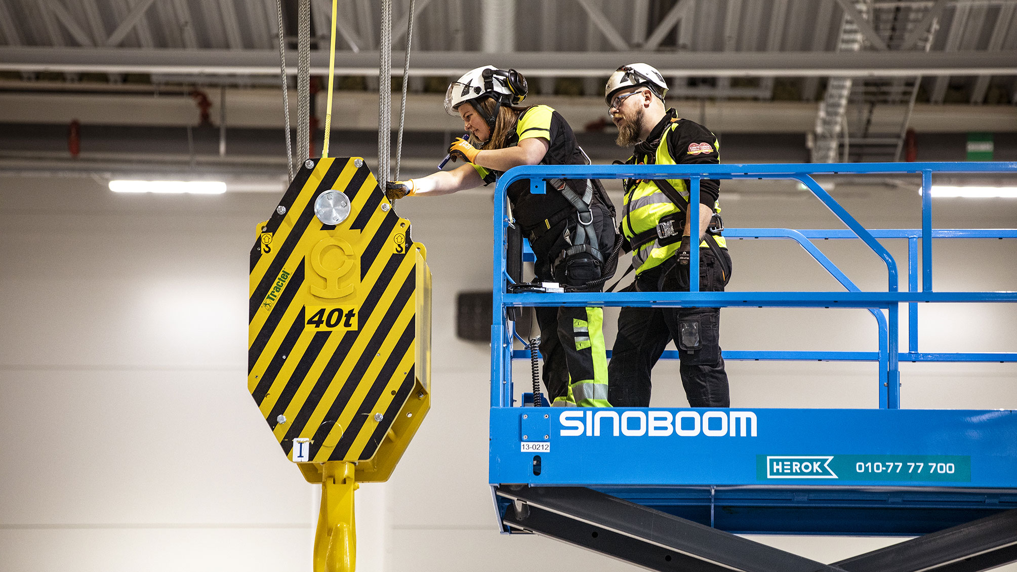 Två besiktningsingenjörer arbetar på en blå arbetsplattform och inspekterar en stor gul och svart lyftkrok märkt med "40t" i en industriell miljö.