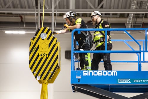 Två besiktningsingenjörer arbetar på en blå arbetsplattform och inspekterar en stor gul och svart lyftkrok märkt med "40t" i en industriell miljö.