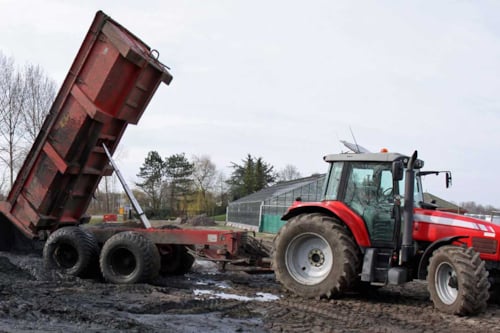 Röd traktor med ett tippande släp på en lerig gård, omgiven av träd och byggnader.
