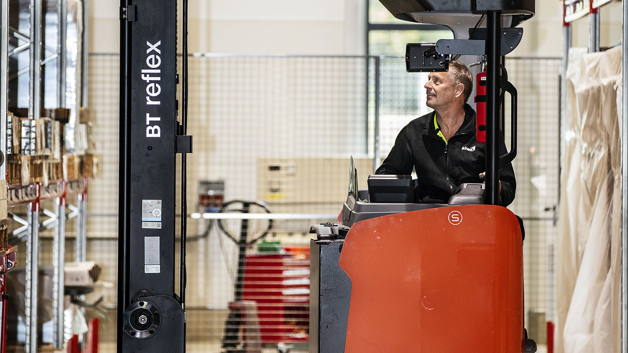 Besiktningsingenjör kontrollerar och kör en orange BT Reflex-styvtruck i en lagerbyggnad med hyllor och utrustning i bakgrunden.
