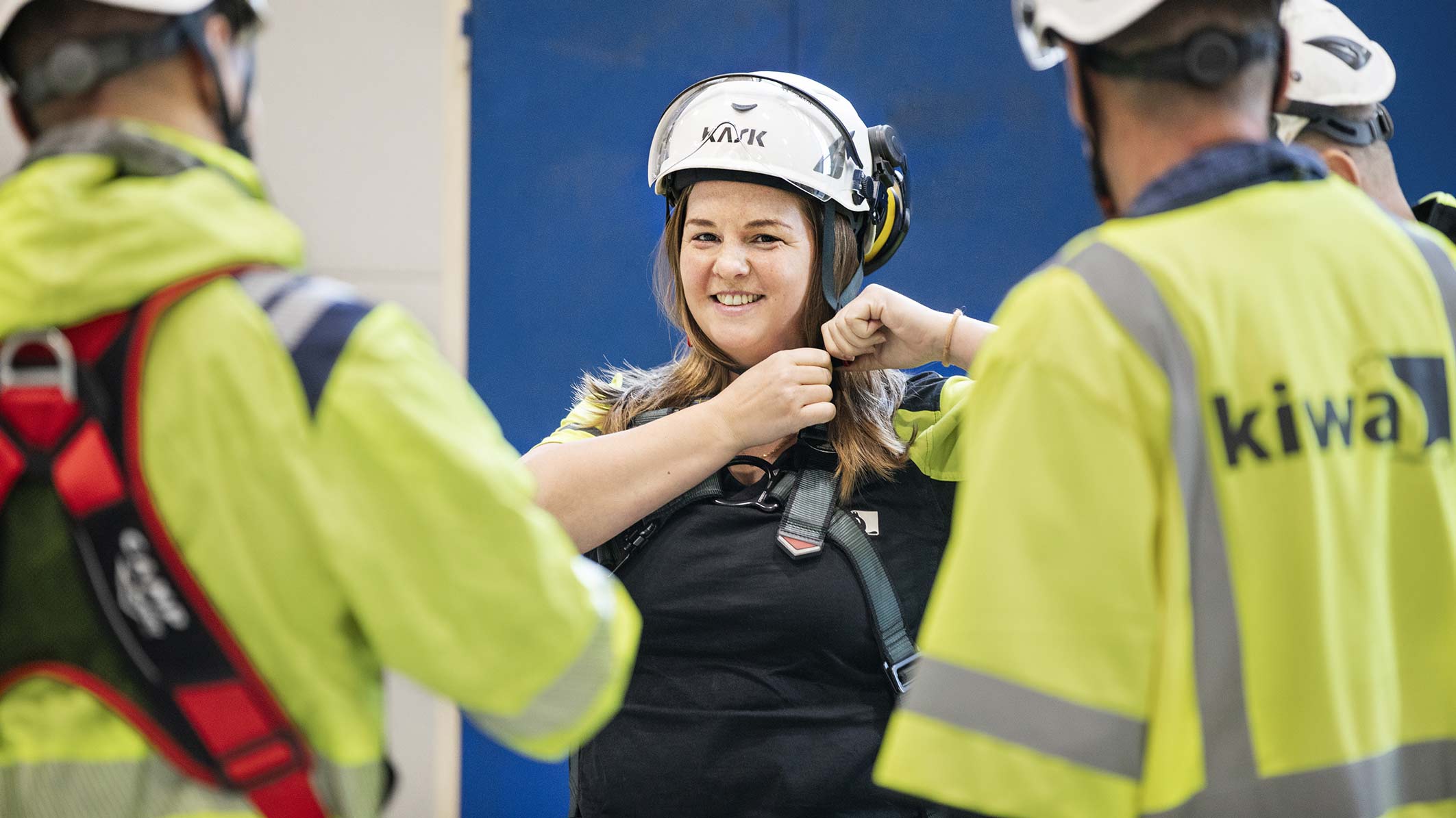 Person i säkerhetsutrustning med hjälm justerar sele, omgiven av två personer i arbetskläder med Kiwa-logotyp.