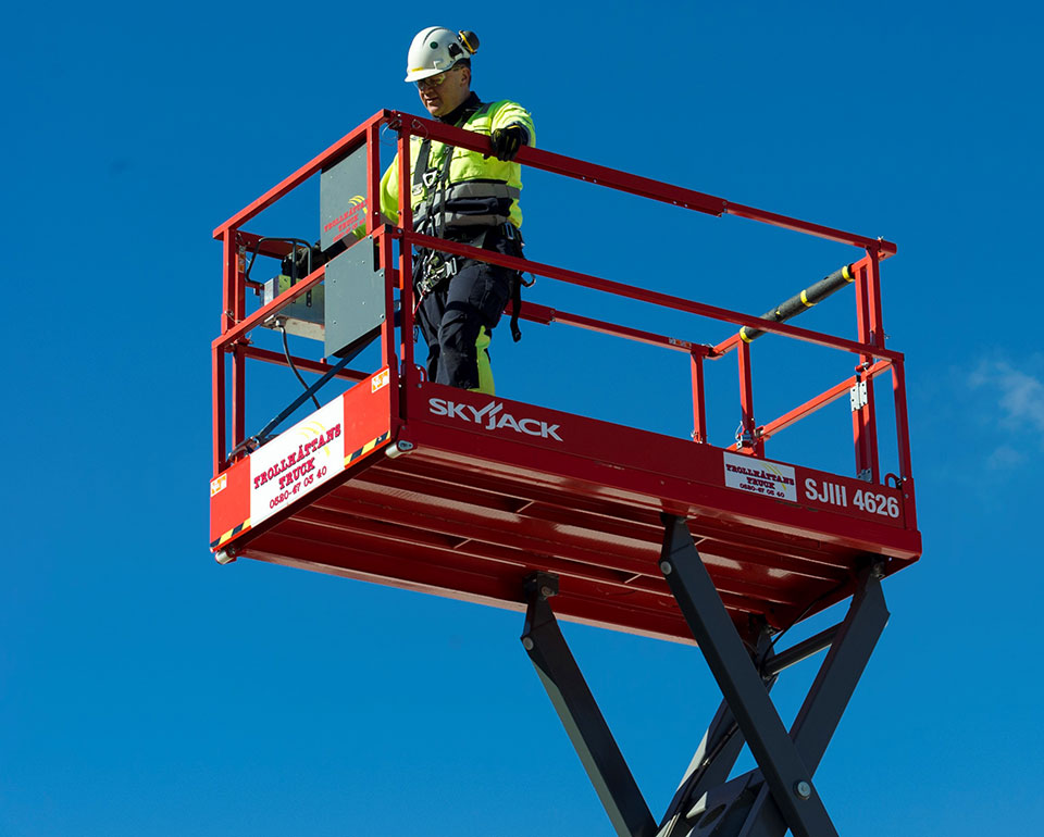 En person står i en röd skylift mot en klarblå himmel, iförd gul skyddsklädsel och säkerhetsutrustning.