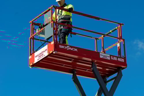 En person står i en röd skylift mot en klarblå himmel, iförd gul skyddsklädsel och säkerhetsutrustning.