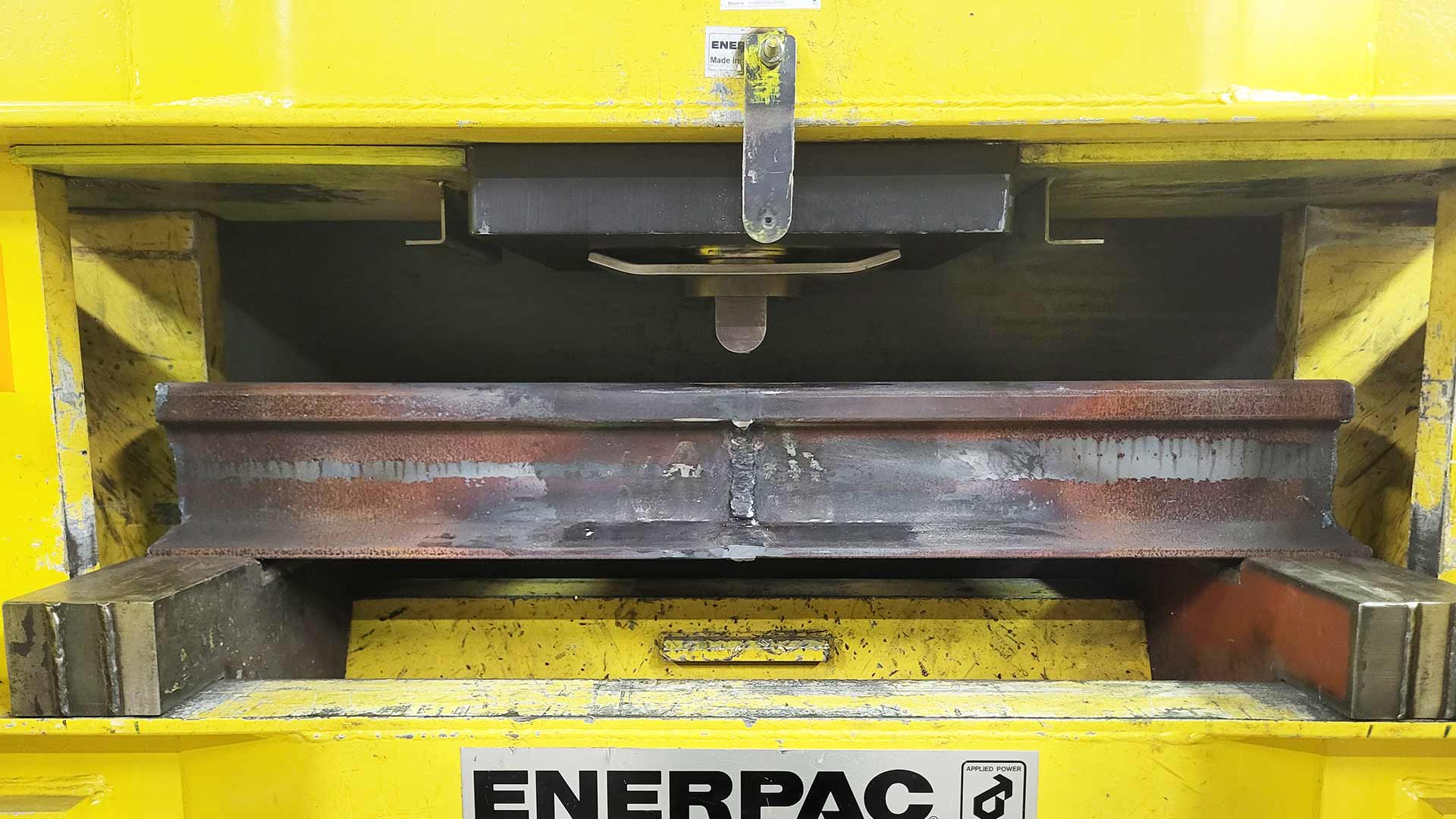 Gul hydraulisk press som böjer en räl i stål, så kallad böjprovning av räl som en metod inom förstörande provning, för att verifiera att rälen uppfyller ställda hållfasthetskrav. 