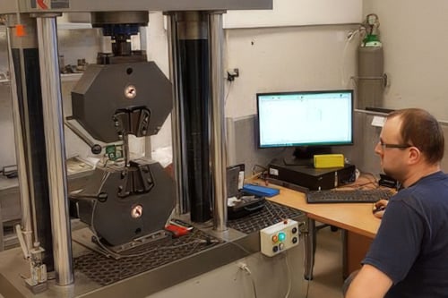 En person sitter nära en materialtestmaskin för ett dragprov i ett laboratorium. En datorskärm och diverse utrustning syns i bakgrunden.
