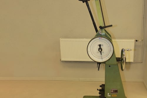 Grön slagprovningsutrustning, med mätare och arm, placerad på ett beige golv framför en ljusgrå vägg.