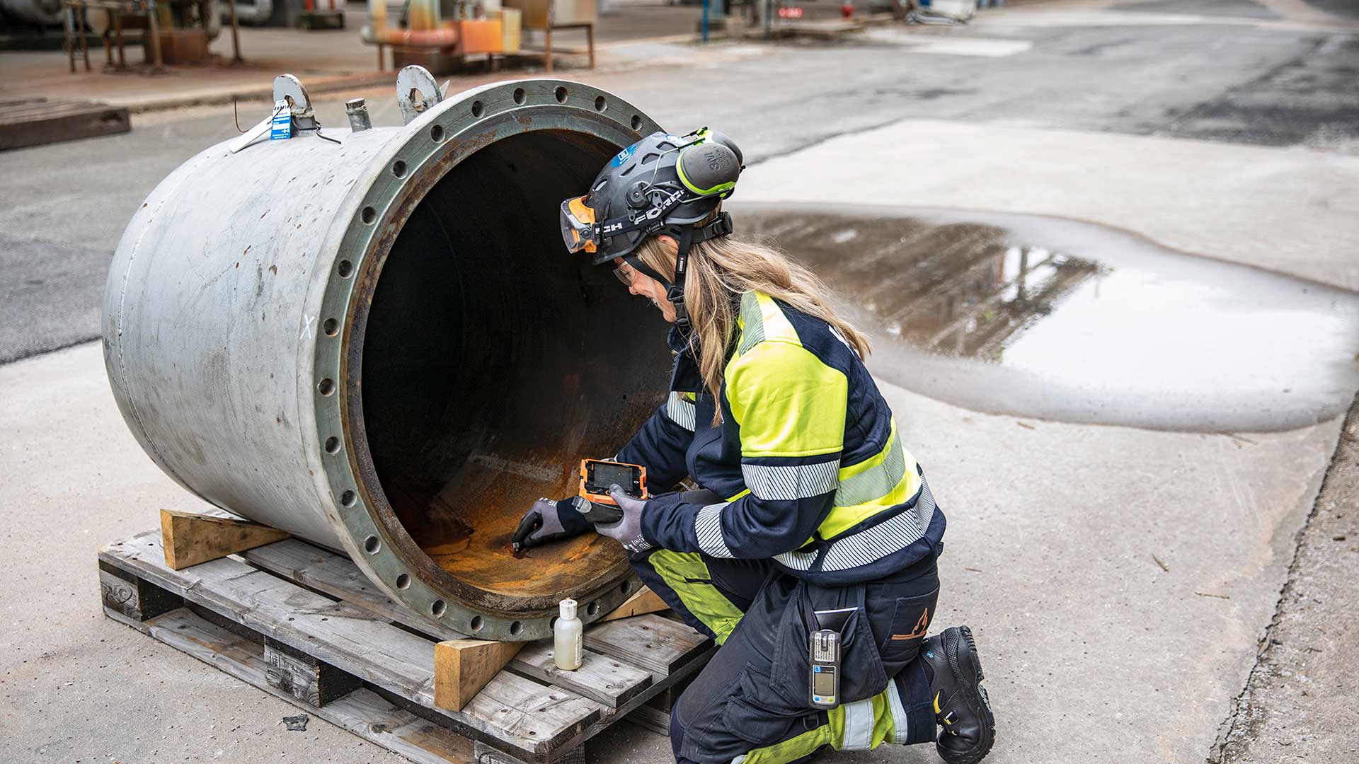 Inspektör i arbetskläder undersöker insidan av ett stort metallrör på en industriplats.