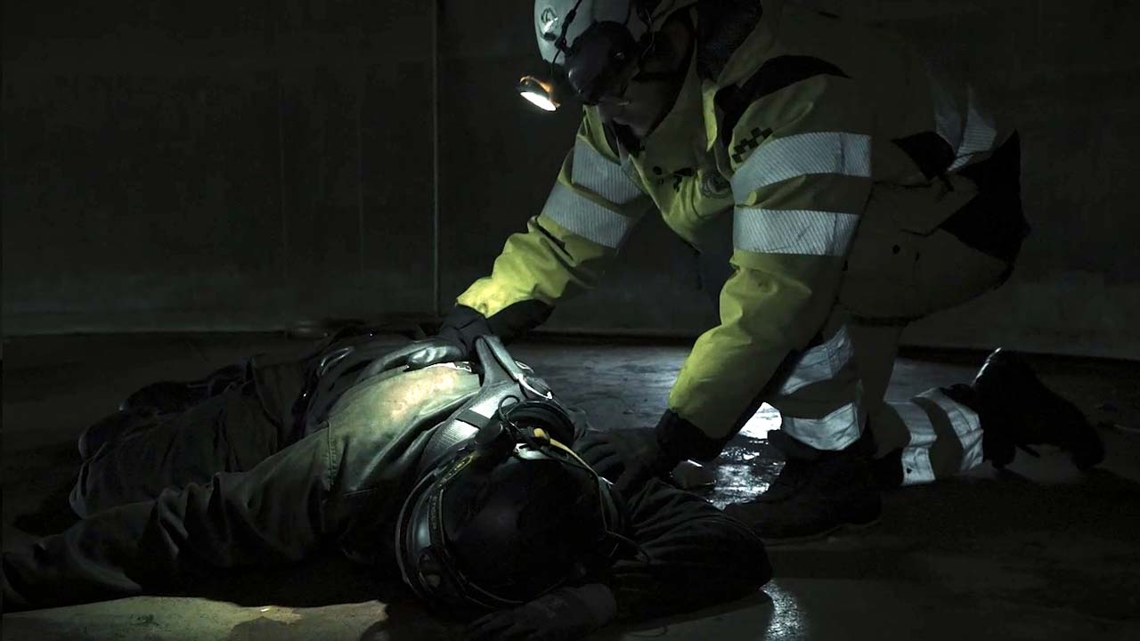 Räddningsarbetare i reflexkläder ger hjälp till en person som ligger på marken i en mörk miljö.