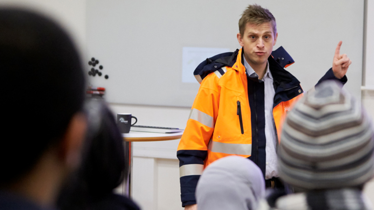 Kiwa trainer in high-visibility orange jacket giving a presentation to an audience in a classroom setting.