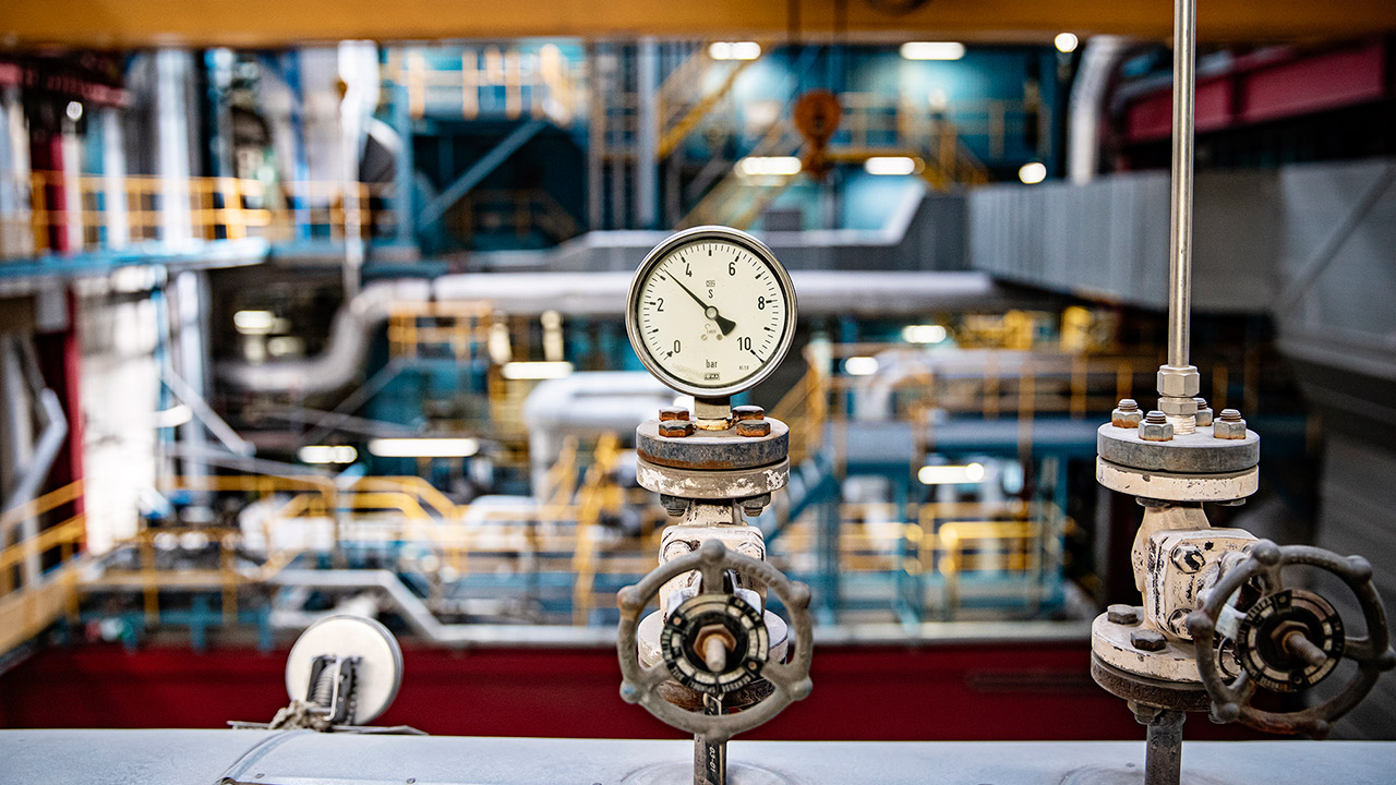 Manometer och ventiler i ett industriellt maskinrum med rör och metallkonstruktioner i bakgrunden.