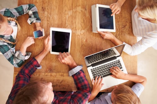 Family of 4 sitting around a table seen from above. All of them using mobile devices like phones, tablets and laptops that have been EMC tested by Kiwa