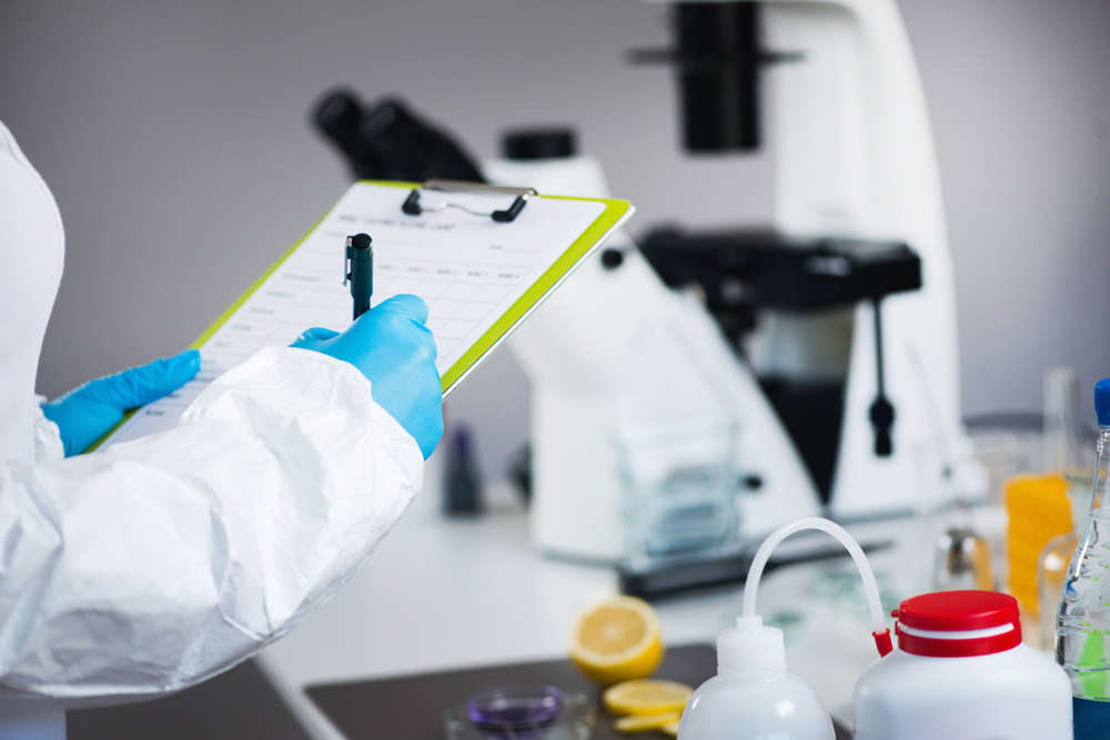 Kiwa lab employee in protective gear reviewing a checklist in a laboratory with a microscope and various equipment in the background.