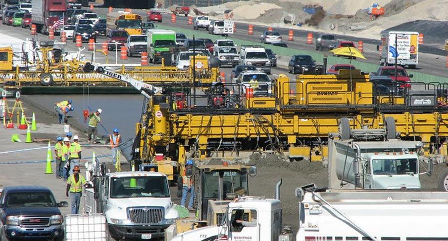 Highway construction site with heavy machinery, workers wearing safety gear, and traffic on adjacent open lanes