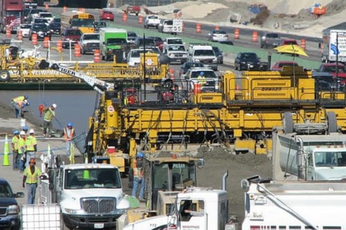 Highway construction site with heavy machinery, workers wearing safety gear, and traffic on adjacent open lanes