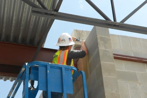 A Kiwa auditor in a safety vest and hard hat using equipment while elevated on a blue lift, working on a concrete block structure with steel beams in the background