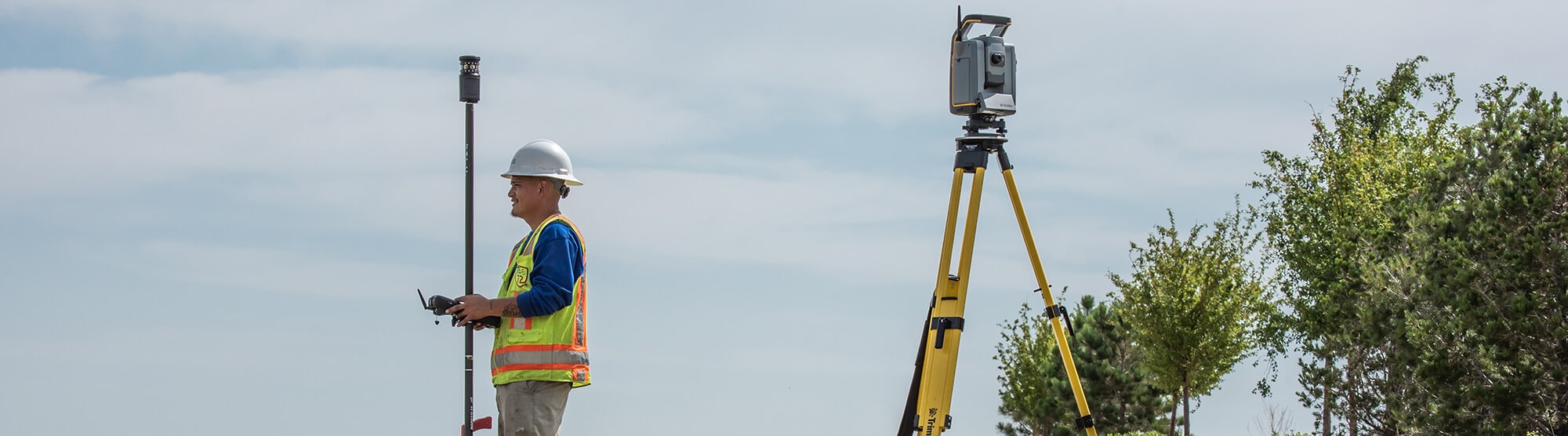 Kiwa T2UE engeneer using a theodolite and rod on a job site, wearing safety gear and helmet, with trees and clear sky in the background