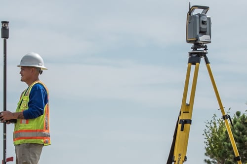 Kiwa T2UE engeneer using a theodolite and rod on a job site, wearing safety gear and helmet, with trees and clear sky in the background