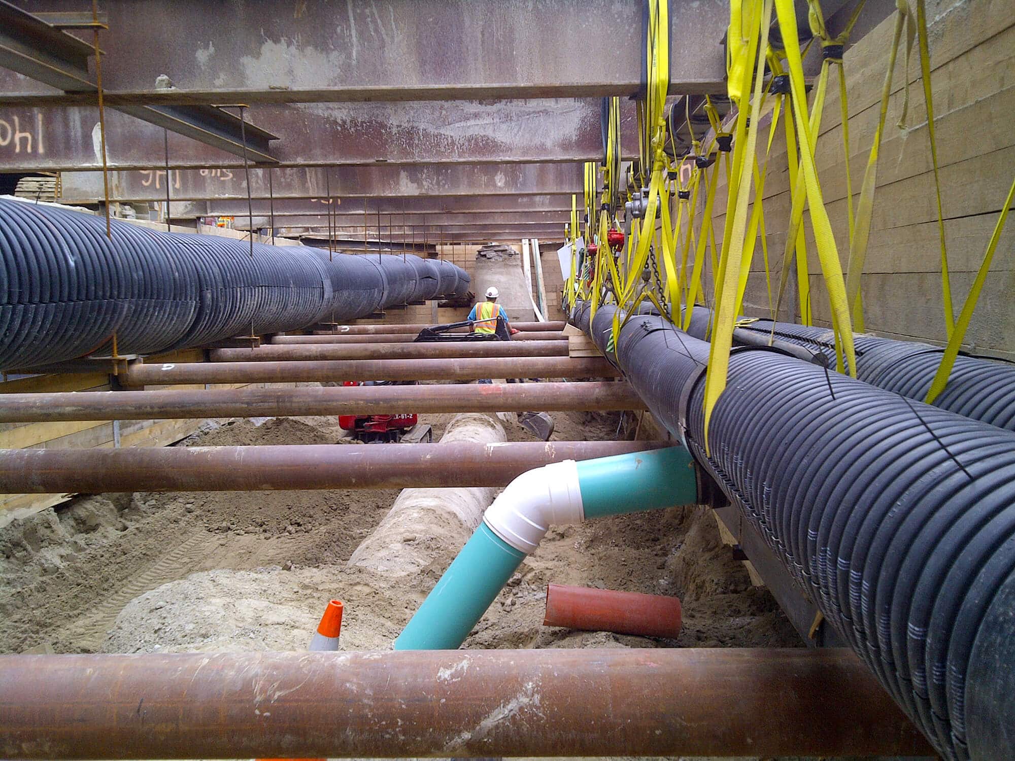 Under-construction underground utility installation site with large black pipes suspended by yellow straps, intersecting blue pipe, and construction equipment in the background