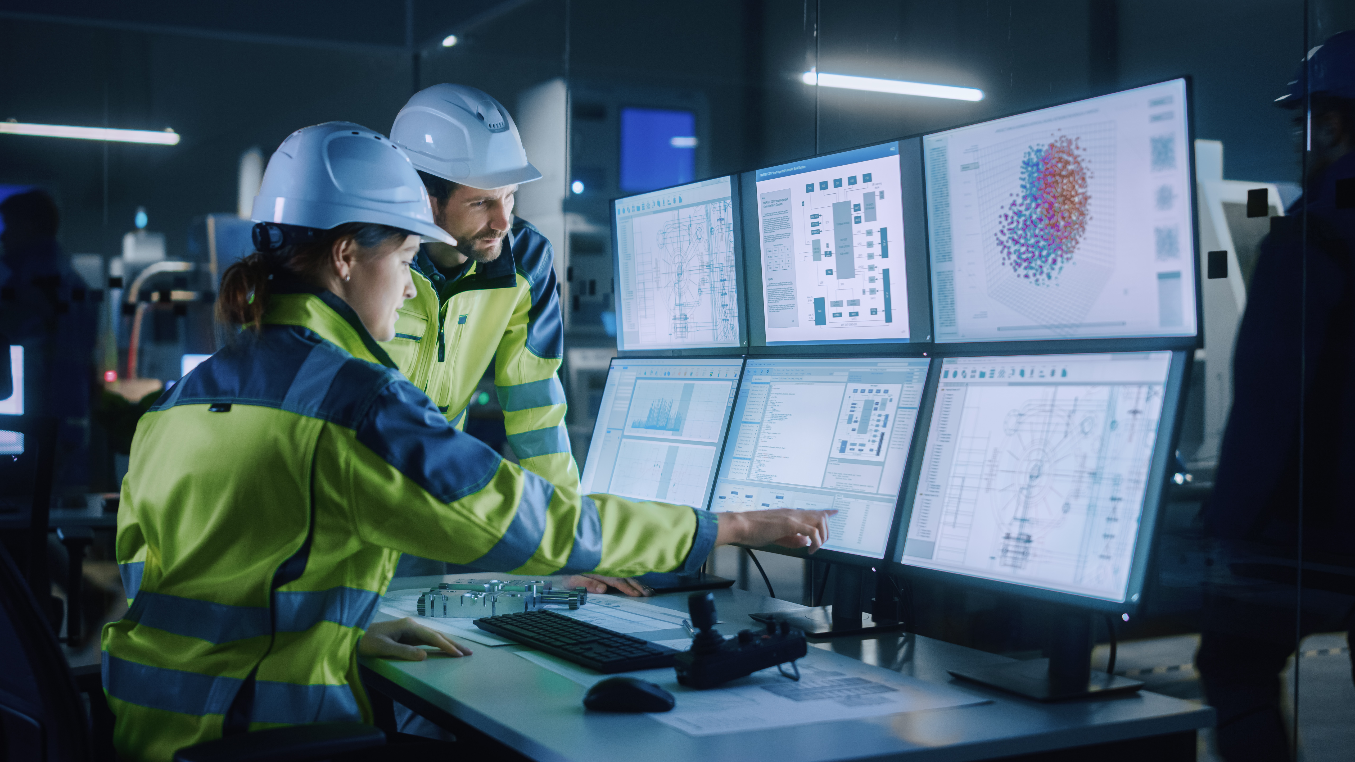 Two Kiwa engineers in hard hats and high-visibility jackets review data on multiple computer monitors in a dimly lit control room.