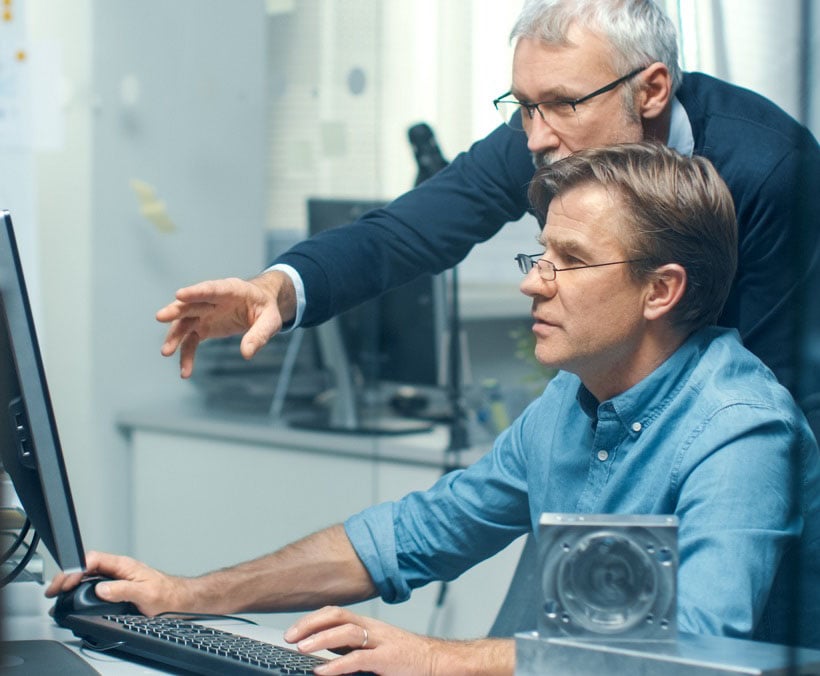 Two men collaborating at a computer in a modern office setting, with one man pointing at the screen, suggesting teamwork and technology-focused work environment.