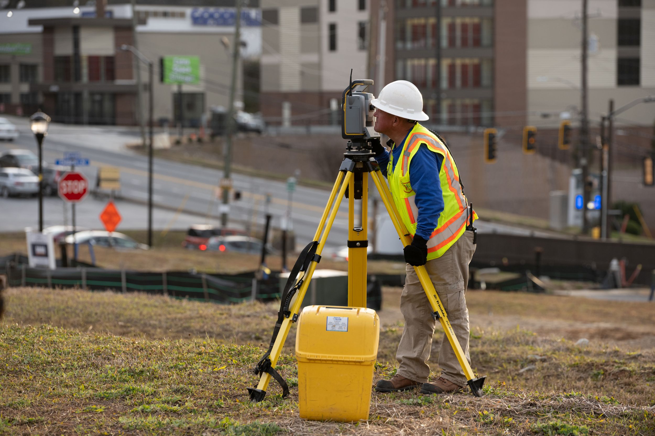 Kiwa employee in a hard hat using a tripod-mounted surveying instrument at a roadside construction site.